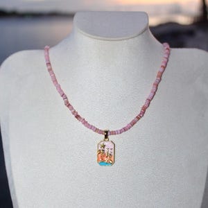 May include: Pink beaded necklace with a rectangular pendant. The pendant has a gold-colored frame and features a colorful illustration of a beach scene. The necklace is displayed on a white mannequin.