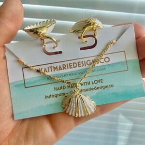 May include: A gold necklace with a white seashell pendant. The necklace is on a white card with the text "kaitmariedesignsco" and "hand made with love" in black. The text "instagram - @kaitmariedesignsco" is also on the card.