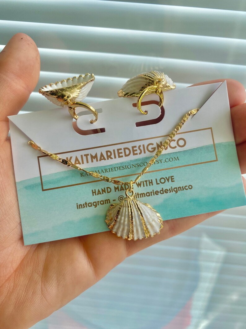 May include: A gold necklace with a white seashell pendant. The necklace is on a white card with the text "kaitmariedesignsco" and "hand made with love" in black. The text "instagram - @kaitmariedesignsco" is also on the card.