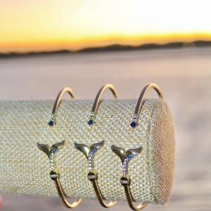 Könnte beinhalten: Drei goldene Armbänder mit einem Wal-Schwanz-Anhänger. Jeder Anhänger ist mit blauen und weißen Kristallen verziert.