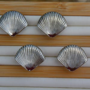 May include: Four silver-colored seashell-shaped buttons on a wooden background. The buttons are arranged in a row, with two on each side of the center.