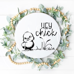 Puede incluir: Un letrero redondo de madera blanco con el texto "Hey Chick" en letras negras. Una ilustración en blanco y negro de un pollito está a la izquierda del texto. El letrero está rodeado de una guirnalda de hojas verdes y cuentas de madera.