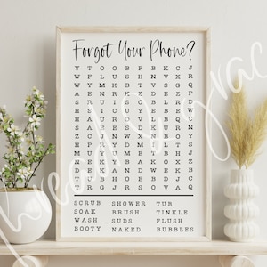 Puede incluir: Una impresi&oacute;n enmarcada con el texto "Forgot Your Phone?" y un crucigrama con las palabras "Scrub, Soak, Wash, Booty, Shower, Brush, Suds, Naked, Tub, Tinkle, Flush, Bubbles" enumeradas debajo. La impresi&oacute;n se muestra en un estante con un jarr&oacute;n blanco y una planta en maceta.