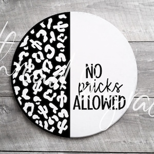 May include: A round wooden sign with a black and white leopard print design on one half and the text "NO PRICKS ALLOWED" on the other half.