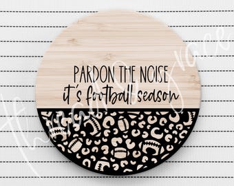 Puede incluir: Un letrero circular de madera con una mitad superior de veta de madera clara con el texto negro "PARDON THE NOISE it's football season" y una mitad inferior negra decorada con un patrón repetitivo de balones de fútbol y manchas de leopardo.