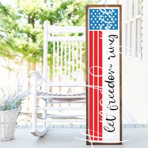 May include: A wooden sign with a red, white, and blue American flag design and the text "Let freedom ring". The sign is leaning against a white rocking chair on a porch.