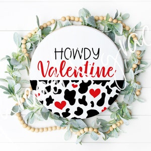 May include: Round wooden sign with the text "Howdy Valentine" in black and red lettering. The bottom half of the sign is decorated with a black and white cow print pattern with red hearts.