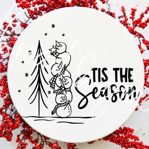 May include: A white circular decorative plate with a black illustration of a Christmas tree and a stack of snowmen wearing scarves. The text "Tis the Season" is written in black script. Red berries surround the plate.