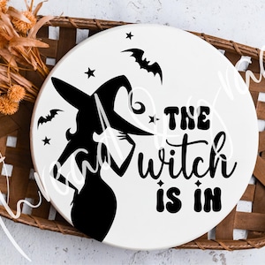 May include: A round, white wooden sign with a black silhouette of a witch wearing a hat, bats, and stars. The text "THE witch IS IN" is in black. The sign is displayed in a brown woven basket, with dried orange flowers.
