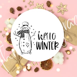 May include: A white round sticker with a black and white illustration of a snowman wearing a striped scarf and a hat. The text "hello winter" is written in black below the snowman.