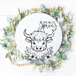 May include: A white round wooden sign with a black and white illustration of a Highland cow with flowers. The sign says "Oh Hello Spring" in black lettering.