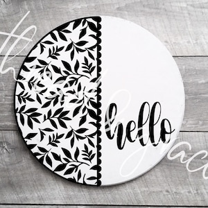 May include: A round, white wooden sign with a black floral design on the left side and the word "hello" in black script on the right. The sign has a decorative scalloped edge.
