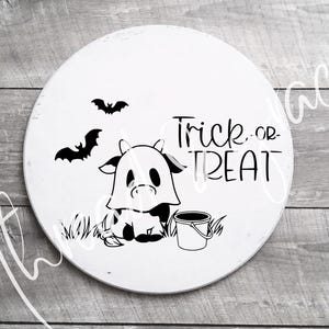 May include: A white round wooden sign with a black and white illustration of a ghost cow holding a bucket. The sign says "Trick or Treat" in black lettering.