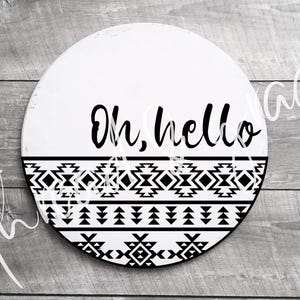 May include: A round, white wooden sign with the words "Oh, hello" in black script. The bottom half features a black and white geometric pattern. The sign is on a gray wooden surface.