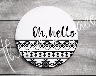 Puede incluir: Un letrero redondo de madera blanca con las palabras "Oh, hello" en escritura negra. La mitad inferior presenta un patrón geométrico en blanco y negro. El letrero está sobre una superficie de madera gris.