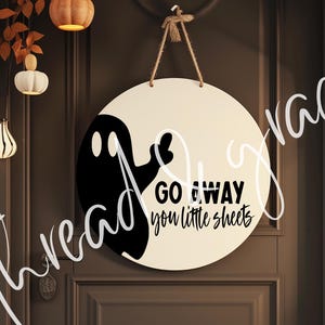 May include: A round, cream-colored wooden sign with a black ghost illustration and the text "GO AWAY you little sheets." The sign is suspended by a natural twine rope. Decorative pumpkins and gourds are visible in the background, suggesting a Halloween theme.