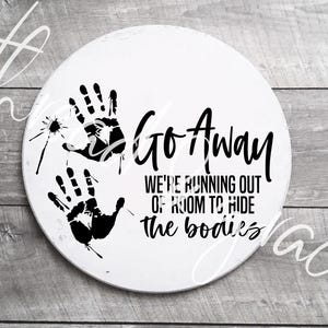 Puede incluir: Cartel decorativo redondo blanco con huellas de manos negras y el texto "Go Away, We're Running Out of Room to Hide the Bodies." El cartel tiene un aspecto envejecido y rústico.