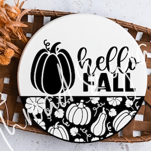 Puede incluir: Un letrero redondo de madera blanca con las palabras "hello FALL" en escritura negra. Un gráfico de calabaza negra está en la parte superior izquierda, y un patrón de calabaza y floral en blanco y negro está en la mitad inferior. El letrero está en una cesta tejida marrón.