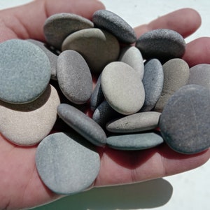 Op de afbeelding: Een verzameling gladde, grijze, bruine en witte stenen in een hand. De stenen zijn van verschillende groottes en vormen.