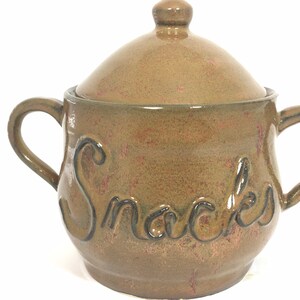 Può includere: Un barattolo per snack in ceramica marrone con coperchio e due manici. La parola "Snacks" è scritta in scuro sulla parte anteriore del barattolo. Il barattolo ha una finitura maculata.
