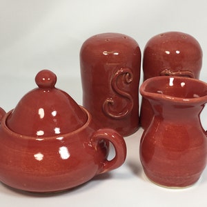 May include: A handmade, red ceramic dining set including a lidded sugar bowl, a creamer pitcher, and two salt and pepper shakers. The salt shaker has an embossed "S". The set is glazed with a glossy finish.