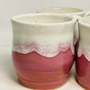 Puede incluir: Cuatro tazas de cerámica hechas a mano. Las tazas tienen un diseño de dos tonos, con una parte superior blanca moteada y una parte inferior de color rosa intenso. La transición entre los colores tiene una apariencia ondulada y texturizada. Las tazas están diseñadas con un asa.