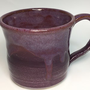 Peut inclure: Une tasse en céramique faite à la main avec un glaçage violet foncé. La tasse a un corps arrondi et une anse incurvée. Le glaçage coule sur les côtés, créant un effet texturé. La tasse mesure environ 10 cm de haut.