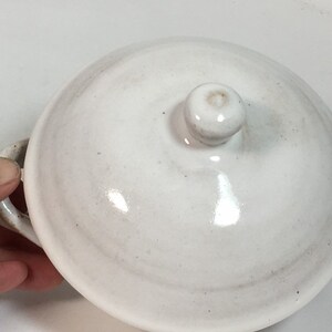 K&ouml;nnte beinhalten: Eine wei&szlig;e Keramiksch&uuml;ssel mit Deckel, zwei Griffen und einem kleinen Knauf oben. Die Sch&uuml;ssel hat eine gl&auml;nzende Oberfl&auml;che und ein leicht gesprenkeltes Aussehen. Die Griffe und der Knauf haben eine hellbraune Farbe.