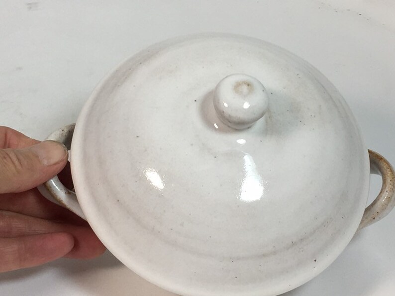 K&ouml;nnte beinhalten: Eine wei&szlig;e Keramiksch&uuml;ssel mit Deckel, zwei Griffen und einem kleinen Knauf oben. Die Sch&uuml;ssel hat eine gl&auml;nzende Oberfl&auml;che und ein leicht gesprenkeltes Aussehen. Die Griffe und der Knauf haben eine hellbraune Farbe.