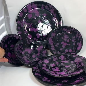 Pode incluir: Um conjunto de louça de cerâmica preta com um padrão roxo salpicado. O conjunto inclui pratos e tigelas de vários tamanhos, perfeitos para servir refeições. O acabamento brilhante realça as vibrantes manchas roxas.