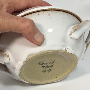 K&ouml;nnte beinhalten: Wei&szlig;er Keramiknapf mit zwei Griffen und Deckel. Die Sch&uuml;ssel hat ein gesprenkeltes, rustikales Aussehen mit braunen Akzenten. Der Boden der Sch&uuml;ssel ist hellbraun und tr&auml;gt den Namen "Gail" und das Datum "7/19".