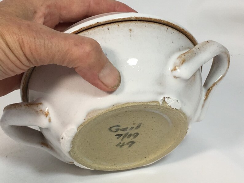 K&ouml;nnte beinhalten: Wei&szlig;er Keramiknapf mit zwei Griffen und Deckel. Die Sch&uuml;ssel hat ein gesprenkeltes, rustikales Aussehen mit braunen Akzenten. Der Boden der Sch&uuml;ssel ist hellbraun und tr&auml;gt den Namen "Gail" und das Datum "7/19".