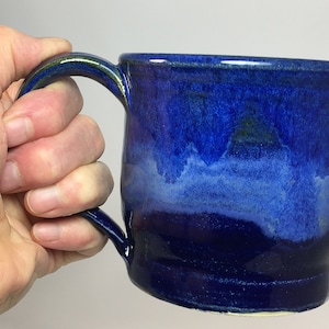 May include: A handmade ceramic mug with a deep blue glaze. The mug has a rounded shape with a handle and a glossy finish. The color transitions from a dark blue at the top to a lighter blue at the bottom.