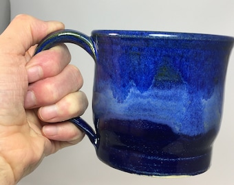 Könnte beinhalten: Eine handgefertigte Keramiktasse mit tiefblauer Glasur. Die Tasse hat eine abgerundete Form mit einem Henkel und einem glänzenden Finish. Die Farbe geht von einem dunklen Blau oben zu einem helleren Blau unten über.