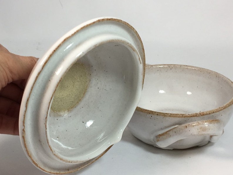 K&ouml;nnte beinhalten: Eine handgefertigte, cremefarbene Keramikschale mit passendem Deckel. Die Schale hat eine abgerundete Form mit einem kleinen Griff an der Seite. Der Deckel hat einen gebogenen Griff. Beide Teile haben eine gesprenkelte Textur und einen braunen Rand.