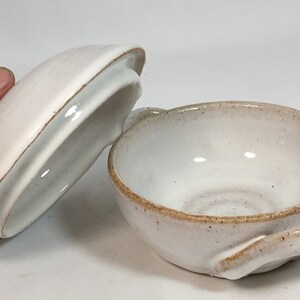 K&ouml;nnte beinhalten: Eine handgefertigte, cremefarbene Keramikschale mit passendem Deckel. Die Schale hat eine runde Form mit einem kleinen Griff und einer gesprenkelten, strukturierten Oberfl&auml;che. Der Deckel ist leicht rosa und hat einen braunen Rand.