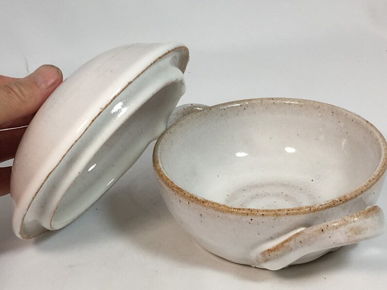 K&ouml;nnte beinhalten: Eine handgefertigte, cremefarbene Keramikschale mit passendem Deckel. Die Schale hat eine runde Form mit einem kleinen Griff und einer gesprenkelten, strukturierten Oberfl&auml;che. Der Deckel ist leicht rosa und hat einen braunen Rand.