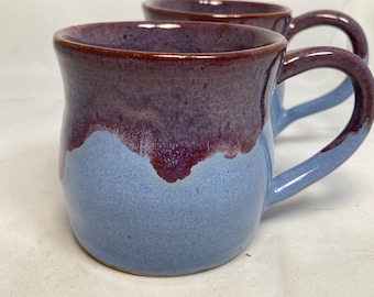 Könnte beinhalten: Zwei handgefertigte Keramikbecher. Die Becher haben eine zweifarbige Glasur, mit einem tiefvioletten Rand, der in einen hellblauen Körper übergeht. Die Griffe sind ebenfalls hellblau. Die Becher sind für Heißgetränke konzipiert.