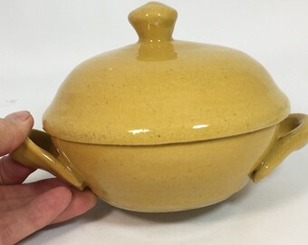 Könnte beinhalten: Eine gelbe Keramik-Servierschüssel mit Deckel und zwei Griffen. Die Schüssel hat eine runde Form mit einem Deckel und einem kleinen Knopf oben. Die Schüssel hat die Farbe Senfgelb.