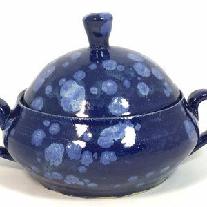 May include: A ceramic sugar bowl with a lid, featuring a deep blue glaze with lighter blue speckles. The bowl has two loop handles and a small knob on the lid. A decorative kitchenware item.