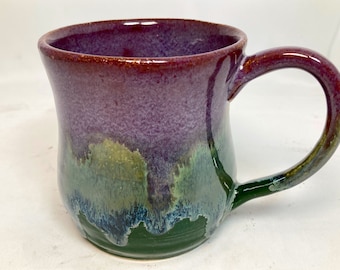 Könnte beinhalten: Eine handgefertigte Keramik-Tasse mit glänzendem Finish. Die Tasse hat einen Farbverlauf, der von einem tiefen burgunderfarbenen Rand zu einem violetten Körper und dann zu Grün und Blau am Boden übergeht. Der Griff ist ebenfalls burgunderfarben.