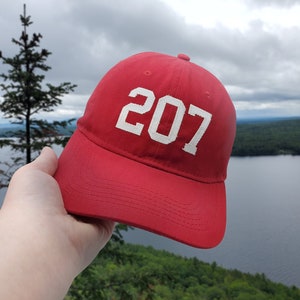 Op de afbeelding: Een rode baseballpet met een wit geborduurd nummer "207" op de voorkant. De pet wordt vastgehouden door een hand voor een achtergrond van een meer en bos.