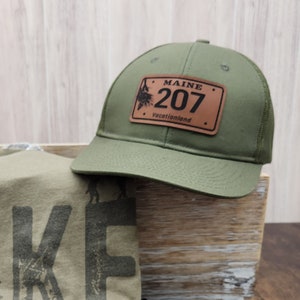 May include: A green t-shirt with the word "HIKE" printed on it in black with a tree graphic in each letter. A green trucker hat with a brown leather patch that says "MAINE 207 vacationland". A brown canvas zippered pouch with a small brown leather patch with a tree graphic and the words "ROUGE PINE" printed on it.