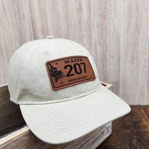 May include: A beige baseball cap with a brown leather patch that says "MAINE 207 Vacationland" with a pine tree design.