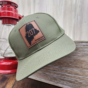 May include: A green trucker hat with a brown leather patch featuring the outline of the state of Maine and the number 207. The patch also includes the text "Rogue Life Maine".