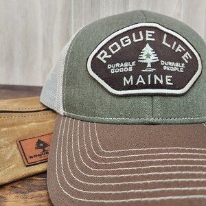 May include: A green and brown baseball cap with a white mesh back. The cap features a brown patch with the text "Rogue Life" in white, a pine tree graphic, and the text "Durable Goods" and "Durable People" in white. The patch also has the text "Maine" in white.