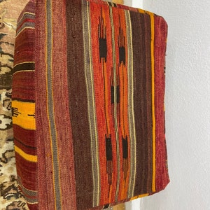 May include: Rectangular floor cushion with a woven, striped pattern in shades of red, orange, brown, and yellow. The cushion has a soft, textured appearance and is likely made of natural fibers. The design features vertical stripes with geometric accents.