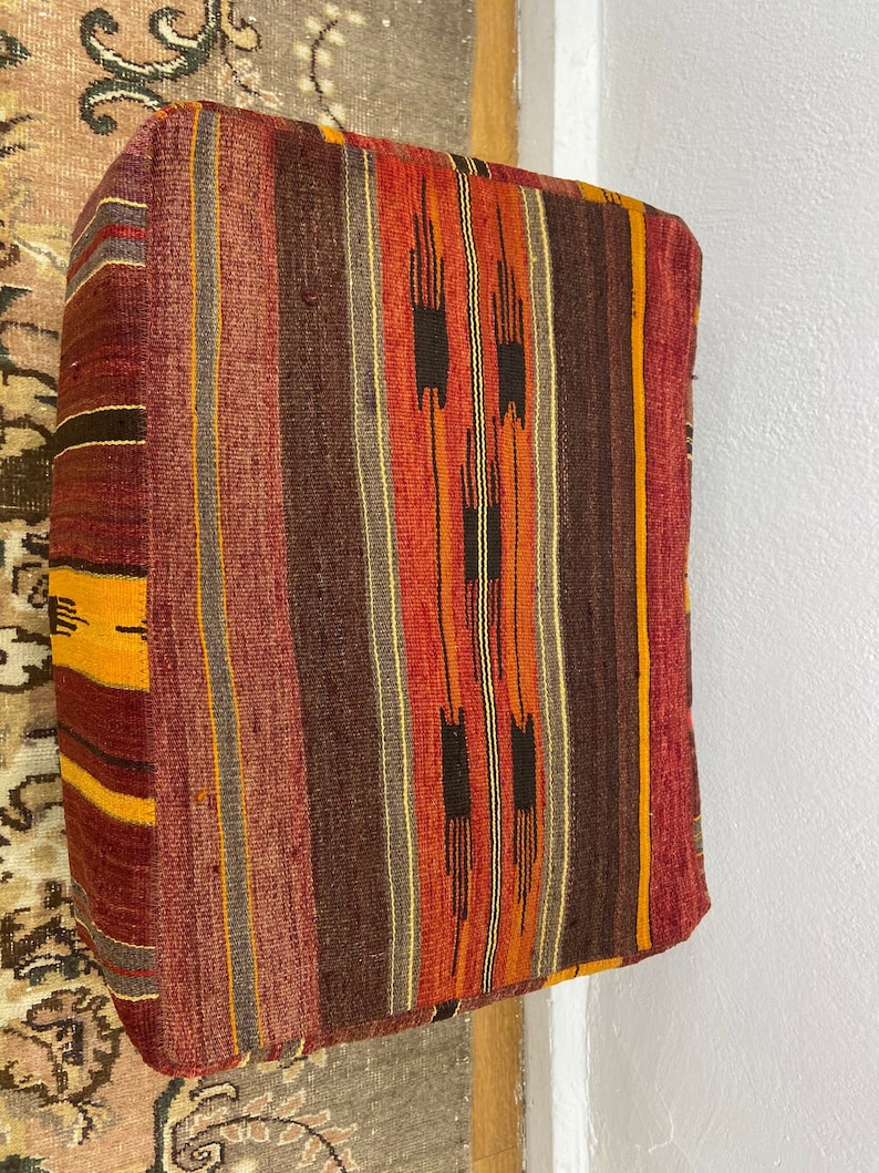 May include: Rectangular floor cushion with a woven, striped pattern in shades of red, orange, brown, and yellow. The cushion has a soft, textured appearance and is likely made of natural fibers. The design features vertical stripes with geometric accents.
