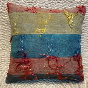 Könnte beinhalten: Ein gestreiftes Kissen mit Patchwork-Muster in Rot-, Gelb-, Blau- und Brauntönen. Das Kissen ist aus einem strukturierten Stoff mit ausgefransten Kanten.