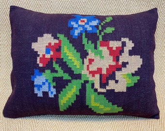 Puede incluir: Un cojín decorativo rectangular con un fondo morado oscuro. El cojín presenta un diseño floral colorido en rojo, azul, verde y beige. Las flores están bordadas en un estilo pixelado, creando una apariencia texturizada.